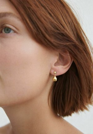 Rövid vörös hajú nő kis arany csepp alakú fülbevalóval, semleges háttér előtt.