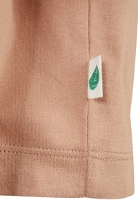 Cotton T-Shirt in sanften Pfirsichtönen mit einem kleinen weißen Etikett im Design eines grünen Blattes, sichtbaren Nähten und einer glatten Textur.