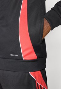 Chaqueta deportiva negra con un destacado panel lateral rojo, detalle de ribete blanco y un logo "AEROREADY" en el dobladillo. Tejido texturizado.