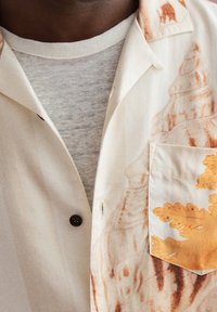 Close-up van een persoon die gelaagde witte en gebroken witte shirts draagt met oranje bladeren en schelppatronen op de shirtzak en kraag.