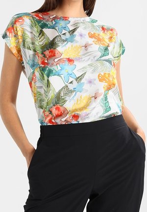 Mujer que lleva una camiseta blanca de manga corta con patrones florales y de hojas coloridos, combinada con pantalones negros de talle alto con las manos en los bolsillos.