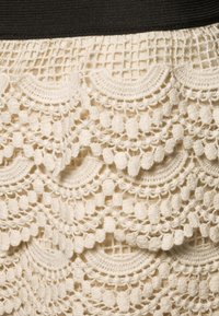 Spetskjol med ett texturerat virkadesign i krämfärg, med scalloped mönster och en svart elastisk midjeband.