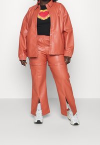 Giacca oversize in similpelle color corallo e pantaloni abbinati con spacchi laterali; t-shirt nera con design di cuore ricamato; sneakers bianche.