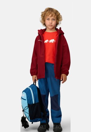 Röd softshelljacka, orange t-shirt med vitt elefanttryck, blå byxor, svarta skor, håller en blå och svart ryggsäck med mesh.