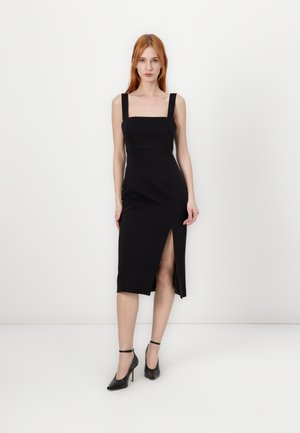 Black sleeveless dress with a square neckline and side slit, made of smooth fabric. Paired with black high-heeled shoes.