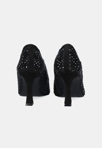 Zapatos de tacón alto negros con un exterior texturizado y tachonado, una punta de zapato lisa y un tacón delgado y cónico. La vista trasera muestra los detalles del diseño.