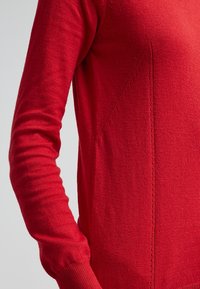Roter Strickpullover mit langen Ärmeln, der ein enges Design, einen strukturierten Stoff und diagonale Nahtdetails am Körper aufweist.