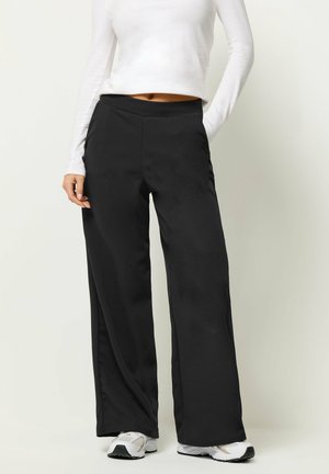 Kobieta ubrana w czarne spodnie szerokie nogawki, biały krótki top z długim rękawem i białe tenisówki, stojąca na jasnym tle.