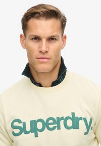 Sudadera crema con el logo verde de "Superdry", superpuesta sobre una camisa de franela azul oscuro con un patrón de cuadro y cuello visible.