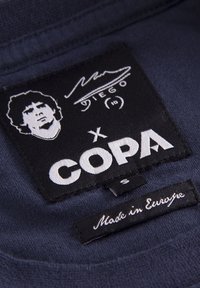 Etiqueta de tela negra con impresión blanca, que presenta un retrato estilizado, una firma y el logo "COPA"; la pequeña etiqueta indica "Hecho en Europa."