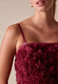 Bordeauxrode jurk met een bloemenoverlay van rozenvormige stof, smalle bandjes en een aansluitende lijfjesstructuur. Subtiele glans op het materiaal.