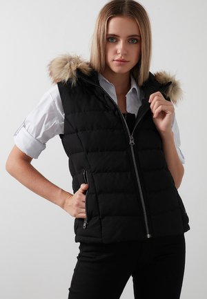 Sort puffervest med fake fur hætte, lynlås foran, sidelommer og kviltet tekstur. Båret over en hvid skjorte med oprullede ærmer.