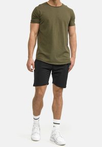 Camiseta de manga corta verde oliva combinada con pantalones cortos negros. Zapatillas de deporte blancas con detalles negros y calcetas con rayas negras en la parte superior.