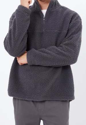 Osoba nosząca ciemnoszary, teksturowany polarowy pulower z kołnierzem na pół zamek, ramiona skrzyżowane, ręka dotykająca brody, na jednolitym tle.