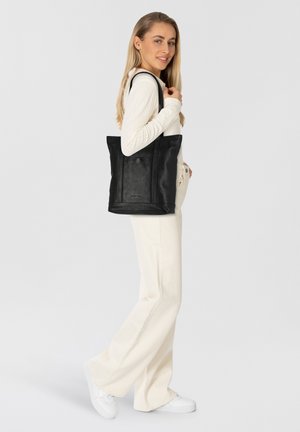 Zwarte leren tote bag met twee handvatten, voorzien van een minimalistisch ontwerp en een gladde textuur. Model staat in een casual witte outfit.