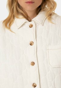 Gep quiltete cremefarbene Jacke mit einem strukturierten Kragen, braunen Knöpfen und einer linken Brusttasche, die ein strukturiertes Rautenmuster aufweist.