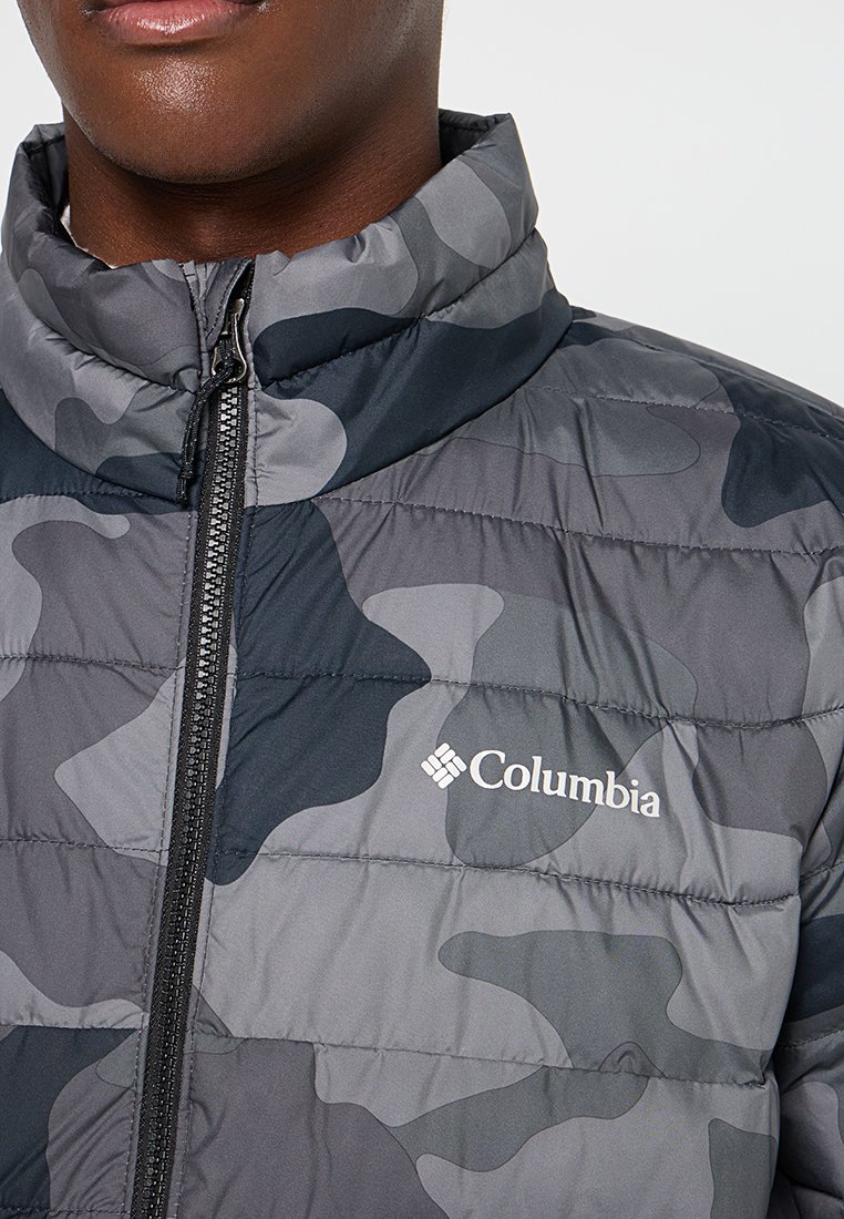 Chaqueta de camuflaje gris con un alto cuello, diseño acolchado y cierre de cremallera. Presenta un logo blanco de Columbia en el pecho. Textura suave.