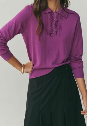 Femme portant un haut à manches longues en maille violette avec des détails à volants et des boutons, assorti à une jupe noire, debout avec une main sur la hanche.