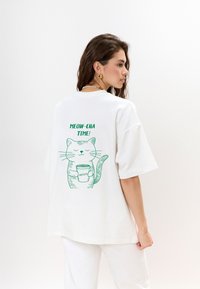 Witte oversized t-shirt met een groene illustratie van een kat die een matcha-beker vasthoudt, met de tekst "MEOW-CHA TIME!" op de achterkant.