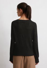 Zwarte langemouwen top van lichtgewicht, doorschijnende stof met een ronde halslijn. Glad textuur, ongeversierde achterkant. Gecombineerd met beige broeken.