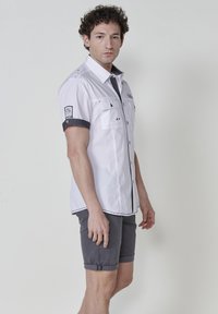 Camisa blanca de manga corta con detalles negros, dos bolsillos en el pecho y puños doblados; combinada con pantalones cortos grises que tienen el dobladillo enrollado.