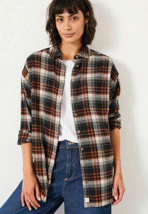 Long-sleeve plaid shirt in black, orange, and beige. Features a button front, rolled sleeves, and a relaxed fit. Worn over a white top.