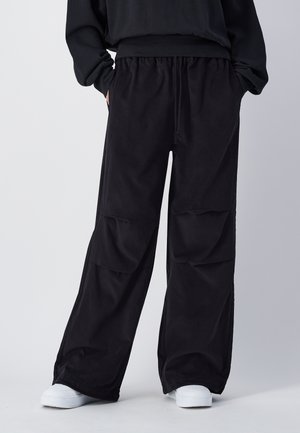 Schwarze weit geschnittene Cargo-Hose mit elastischem Bund, zwei Seitentaschen und einer glatten Stoffoberfläche. Kombiniert mit weißen Sneakers.