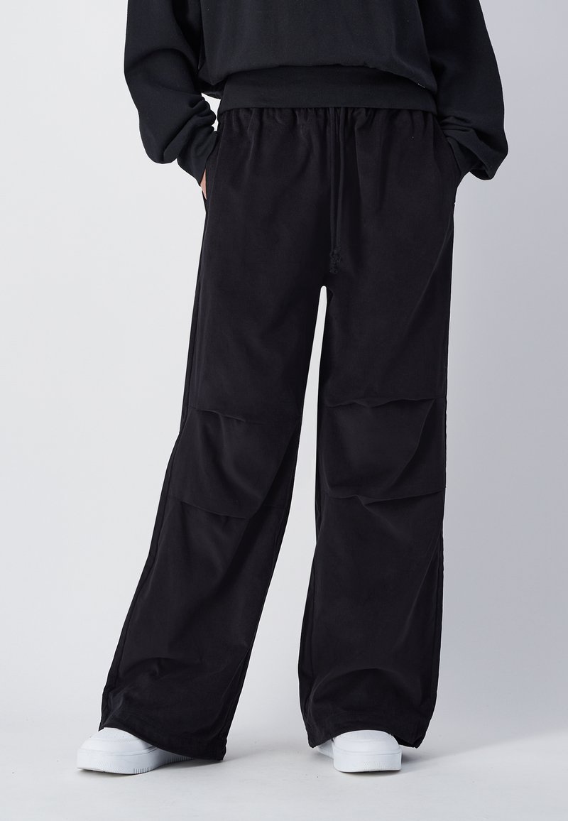 Pantalons cargo larges noirs avec une taille élastique, deux poches latérales et une texture de tissu lisse. Stylés avec des baskets blanches.