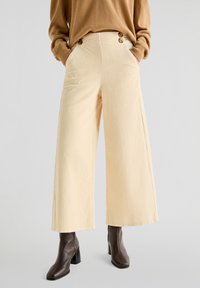 Beige corduroy bukser med brede ben, vertikale striber, høj talje og to frontlommer; har knapdetaljer ved taljen.