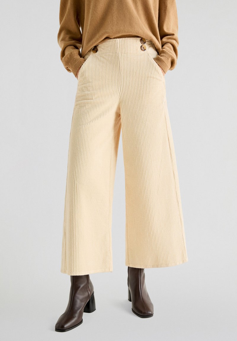 Beige corduroy bukser med brede ben, vertikale striber, høj talje og to frontlommer; har knapdetaljer ved taljen.