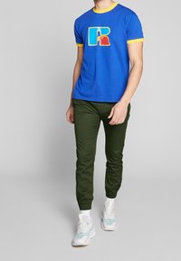 T-shirt en coton bleu avec bordure jaune présentant un motif coloré en forme de "R", associé à un pantalon slim vert et des baskets blanches avec des accents bleus.