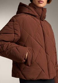 Brun quiltet pufferjakke med høj krave, der har en firkantet pasform og sidelommer. Materialet er glat med en mat finish.