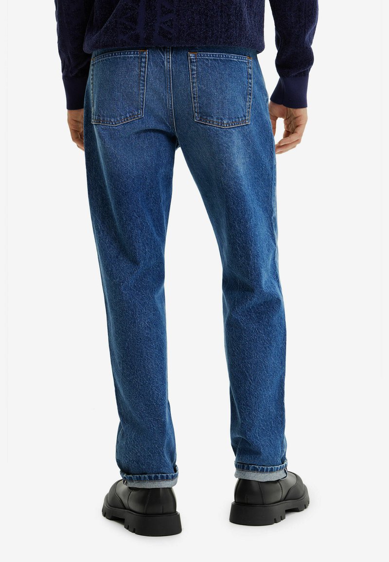 Homme portant un pantalon en jean bleu avec des ourlets retournés, des chaussures noires épaisses et un pull en maille bleu foncé, vu de l'arrière.