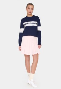 Navy sweater with white "après tennis" text and stripes, paired with a pleated light pink skirt and white sneakers. Cream socks complete the look.