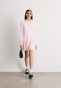 Ružový pletený šaty s golierom, skladanou sukňou, nosené s bielymi ponožkami a čiernymi topánkami. Príslušenstvo tvoria veľké slnečné okuliare a čierna taška.