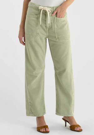 Pantalon large couleur vert clair avec grandes poches plaquées à l'avant et taille à cordon blanc, porté avec des sandales à talons ouvertes marron.