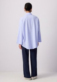 Cămașă supradimensionată, cu dungi verticale albastre deschise, cu mâneci lungi, detalii cu pliuri pe spate, asortată cu pantaloni largi negri și încălțăminte deschisă.