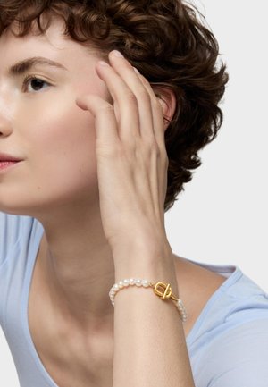 Młoda osoba z krótkimi kręconymi włosami, ubrana w jasnoniebieski top, dotykająca twarzy jedną dłonią ozdobioną perłową bransoletką z złotym zapięciem.