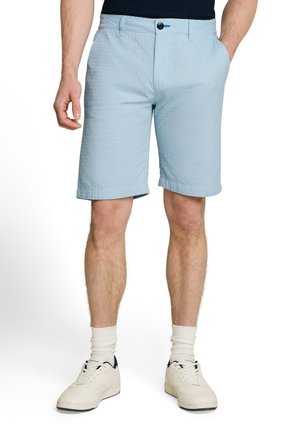 Mann trägt hellblaue, strukturierte Shorts, weiße Socken und weiße Sneakers und steht mit entspannten Händen, eine Hand in der Tasche.