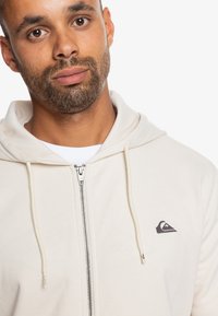 Svetlá béžová mikina na zips s predným zipsom a sťahovacími šnúrkami. Obsahuje nášivku s logom na hrudi a jemnú textilnú štruktúru.