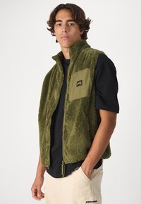 Olive green fleece vest with zip front, high collar, and a chest pocket. Textured fabric and a black logo patch on the pocket.