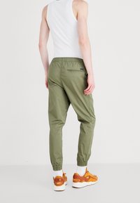 Lätta olivgröna byxor med elastiska muddar, bakficka och dragsko i midjan, tillsammans med orange sneakers på en vit bakgrund.