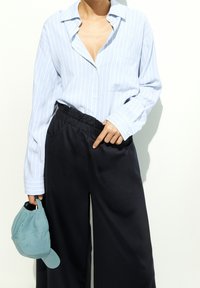 Personne portant une chemise à rayures bleu clair et un pantalon noir à taille élastique, tenant une casquette de baseball turquoise dans la main gauche.