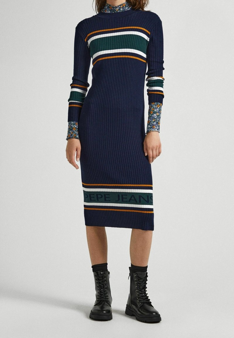Femme portant une robe midi bleu marine côtelée avec des rayures colorées et le texte "PEPE JEANS", accompagnée de manches florales et de bottes noires à lacets.