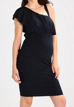 Femme portant une robe noire ajustée à une épaule avec un détail à volants sur la poitrine, debout devant un fond uni.