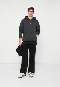Jovem a usar um hoodie e calças pretas da Adidas Originals, sapatilhas brancas da Adidas, segurando uma chapéu de beisebol preto, de pé contra um fundo branco.