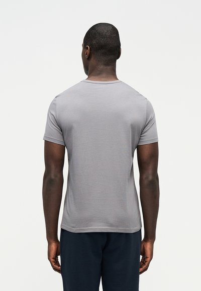 Άνδρας που στέκεται με την πλάτη, φορώντας ανοιχτό γκρι κοντομάνικη μπλούζα και σκούρο παντελόνι σε απλό ανοιχτό φόντο.