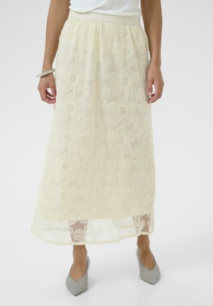 Ženska, ki nosi kremno midi krilo iz cvetlične čipke z bleščicami, belo majico, sive čevlje z ostro konico in srebrno zapestnico na levi roki.