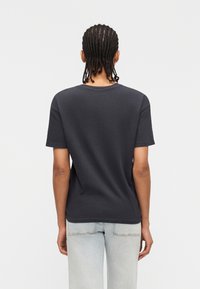 Άτομο με πλεγμένα μαλλιά που φοράει κοντομάνικο μπλουζάκι σκούρου γκρι χρώματος και ανοιχτού μπλε τζιν, στέκεται μπροστά σε ένα απλό λευκό φόντο.