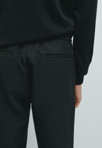 Pantaloni neri con una texture liscia, dotati di due tasche posteriori e una vita elasticizzata, parzialmente mostrati con una maglietta nera.